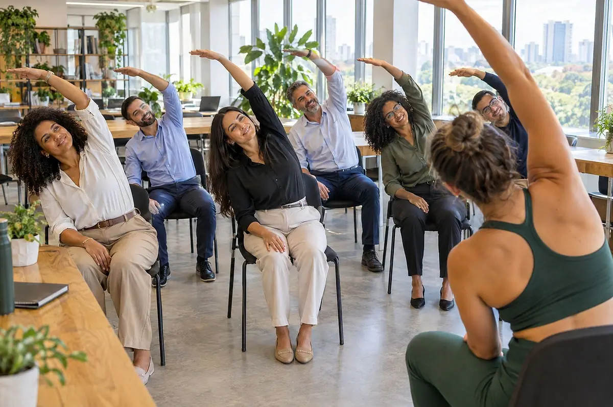 Colaboradores fazendo alongamento lateral sentados em cadeiras, guiados por uma instrutora de yoga em um escritório iluminado