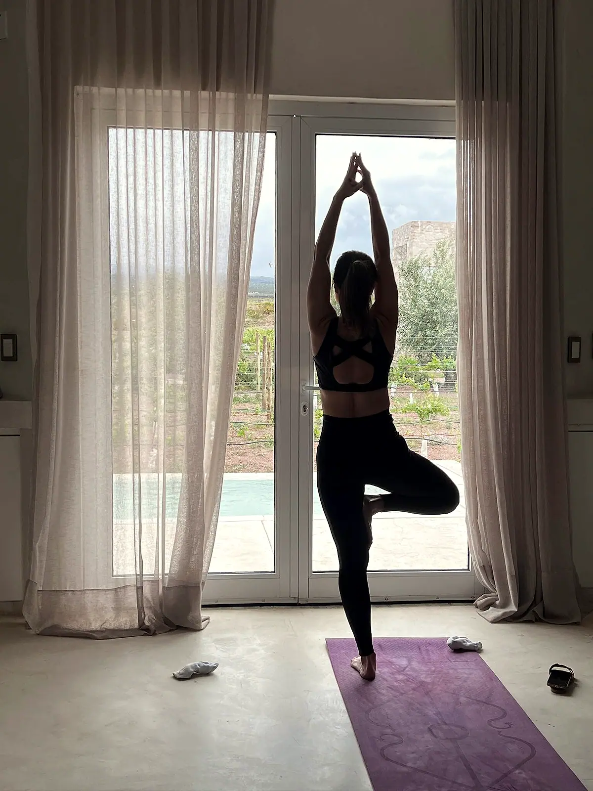 Luciane Lemos na postura da árvore (Vrksasana), mãos unidas acima da cabeça, diante de uma janela com luz natural
