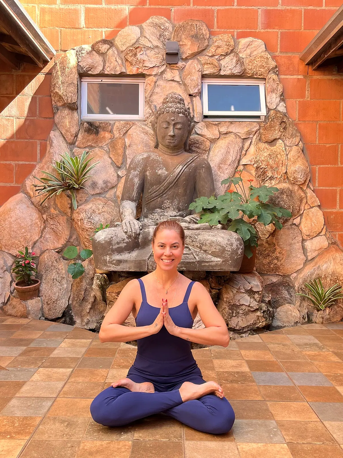 Luciane Lemos sentada em postura de lótus com as mãos em namastê diante de uma estátua de Buda