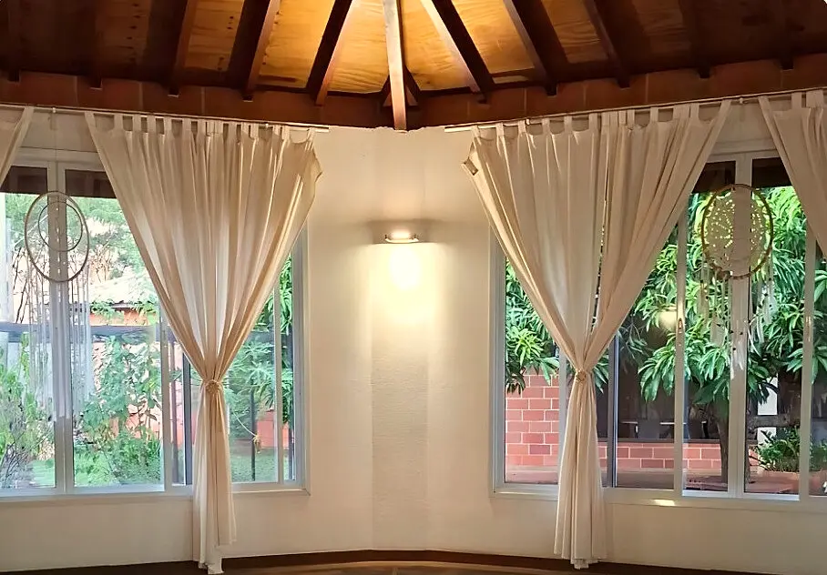 Salão de práticas octogonal com teto de madeira, cortinas claras e grandes janelas